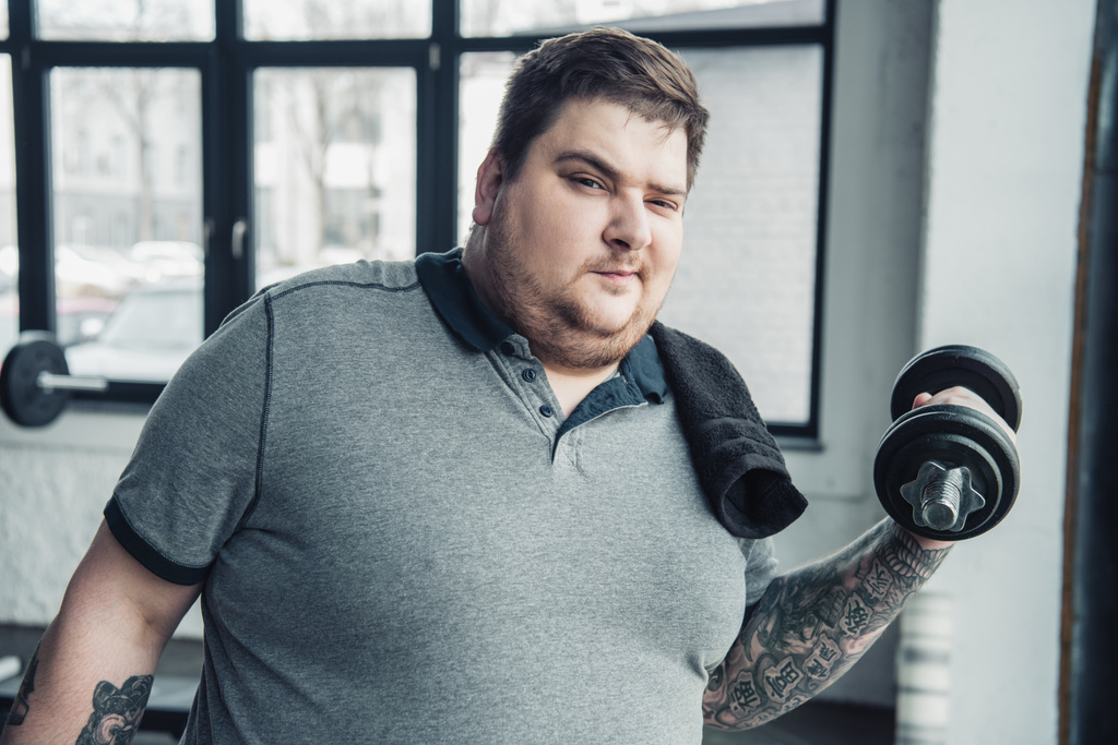 Hombre tatuado con sobrepeso con toalla haciendo ejercicio con mancuerna y mirando a la cámara en el centro deportivo
 - Foto, Imagen