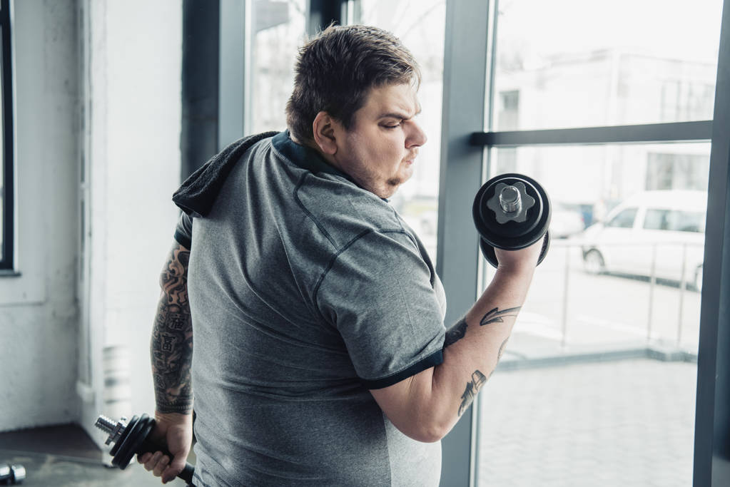 Избыточный вес татуированный человек с полотенцем упражнения с гантелями в спортивном центре
 - Фото, изображение