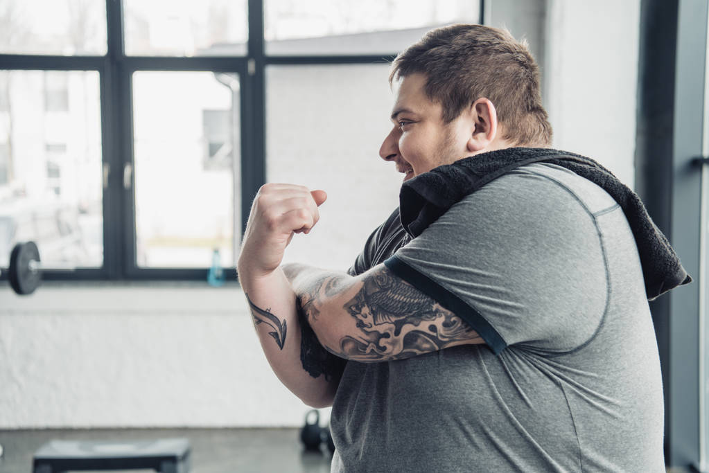 túlsúlyos tetovált ember, fehér póló mosolygó és nyújtás karok a sportközpontban - Fotó, kép