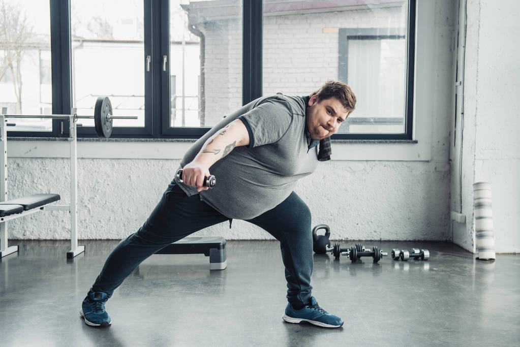 Overgewicht getatoeëerde man in grijs t-shirt met handdoek trainen met halter in sportcentrum - Foto, afbeelding
