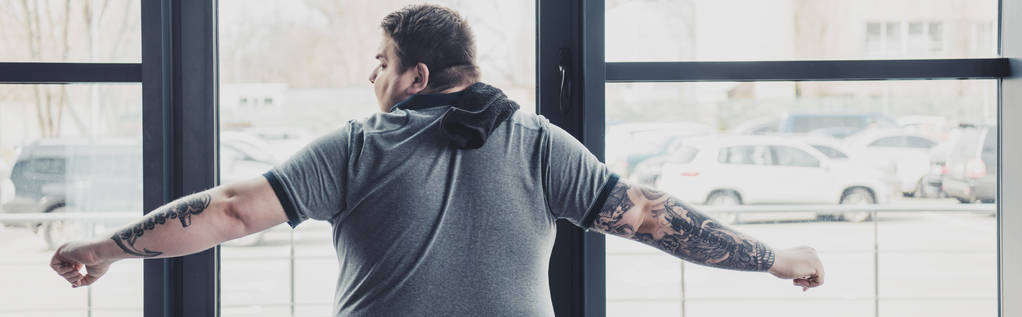 вид ззаду татуйованого чоловіка з надмірною вагою у сірій футболці, що розтягується у спортзалі
 - Фото, зображення