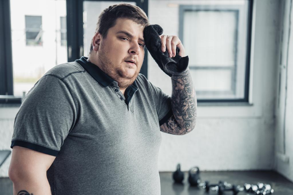 Müder übergewichtiger tätowierter Mann blickt in Kamera und wischt sich im Sportzentrum mit Handtuch das Gesicht ab - Foto, Bild