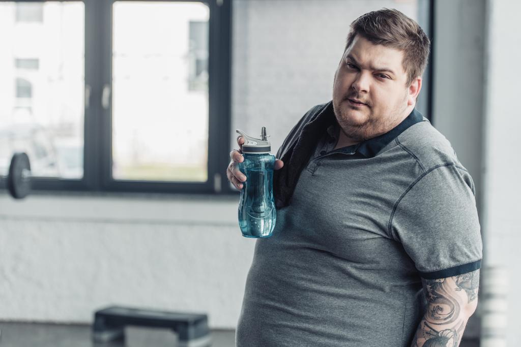 太りすぎの入れ墨の男は、カメラを見て、ジムでスポーツボトルを保持 - 写真・画像