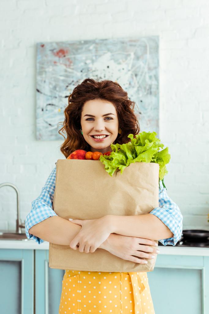 Lachende vrouw in polka dot schort knuffelen papieren zak met rijpe vruchten en groenten - Foto, afbeelding