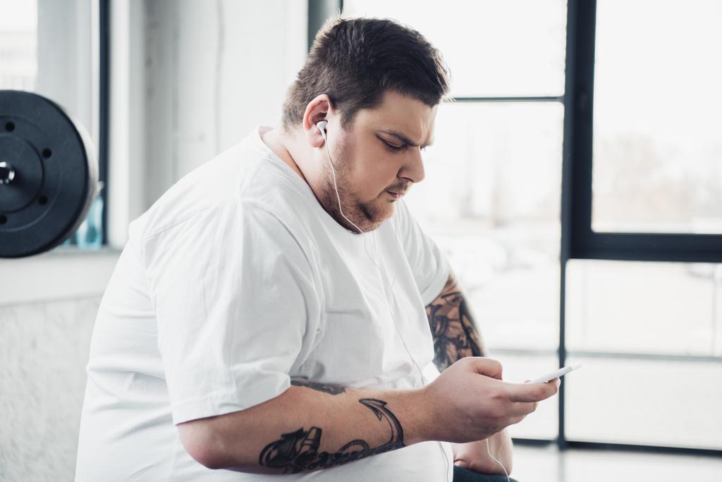 túlsúlyos ember-ban fülhallgató ülő és használ smartphone-on tornaterem - Fotó, kép