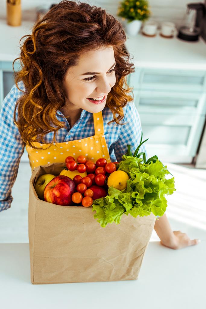 mujer feliz cerca de la bolsa de papel con frutas y verduras maduras en la mesa de la cocina
 - Foto, imagen