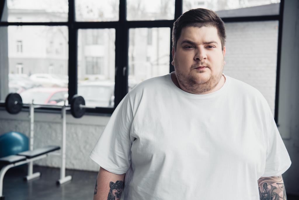 Übergewichtiger, tätowierter Mann in weißem T-Shirt blickt im Sportzentrum mit Kopierraum in die Kamera - Foto, Bild