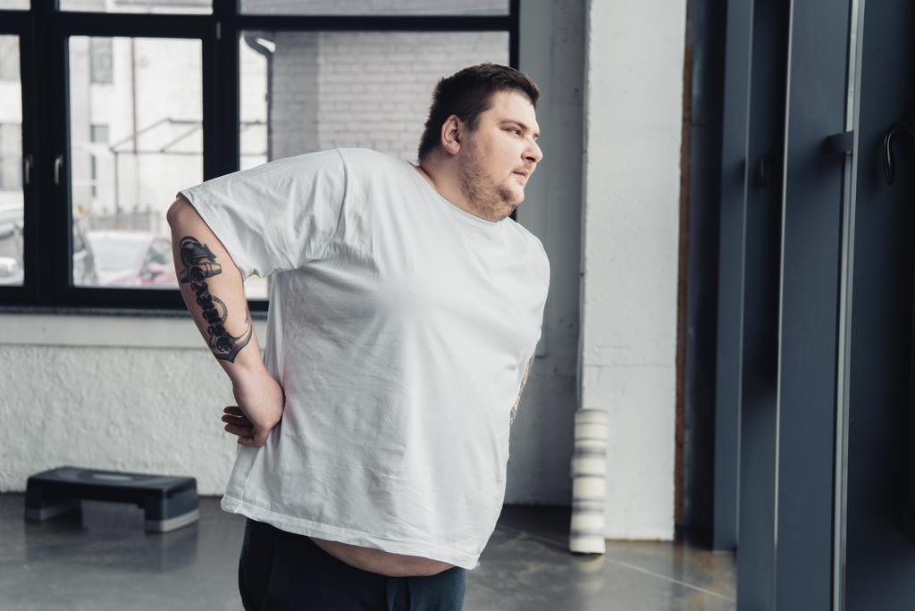 obézní muž v bílém tričku, který se ve sportovním centru protahuje cvičením - Fotografie, Obrázek