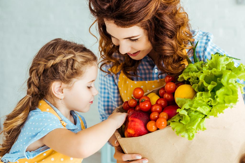 glückliche Mutter zeigt Tochter Papiertüte mit reifem Obst und Gemüse - Foto, Bild