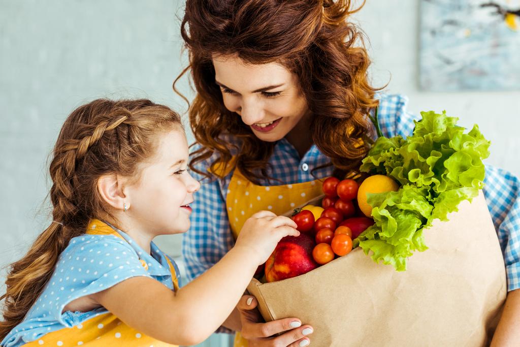 boldog anya mutatja mosolygó lánya táska a papír zsák érett gyümölcs és zöldség - Fotó, kép