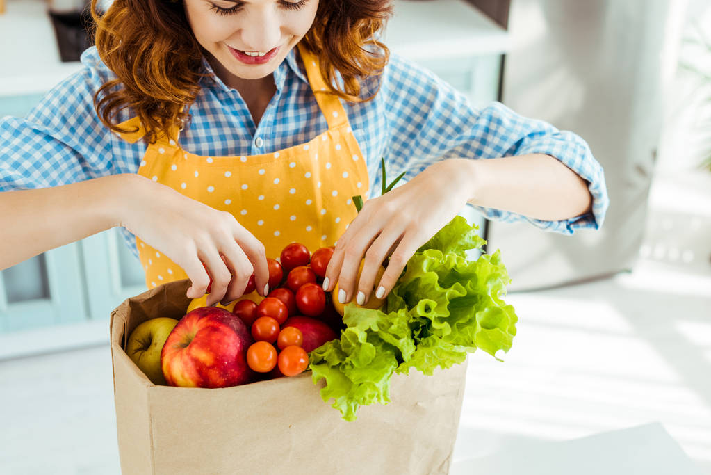 happy woman in polka dot yellow apron taking fruits and vegetables out of paper bag - 写真・画像