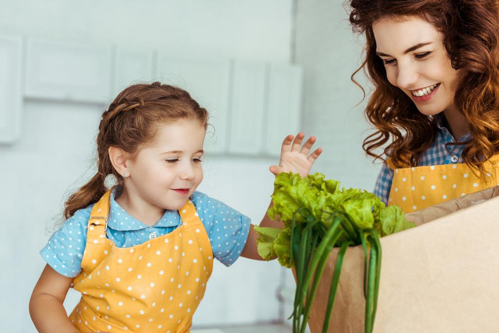 happy mother in polka dot apron holding paper bag with lettuce near smiling daughter - Photo, Image