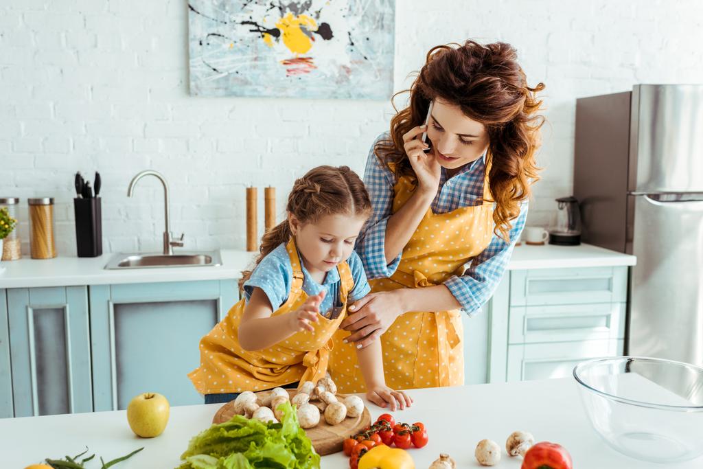 mãe falando no smartphone e olhando para a filha de pé perto da mesa com legumes
 - Foto, Imagem