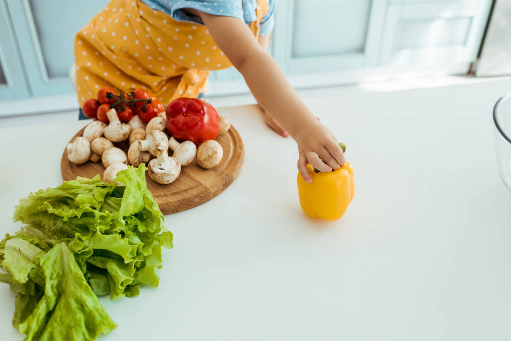 cropped view of child in polka dot apron holding yellow bell pepper on table with vegetables and chopping board - Photo, Image