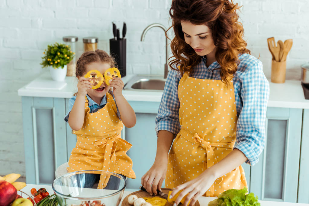 bonito filha segurando sino pimenta fatias na frente dos olhos enquanto mãe corte legumes
 - Foto, Imagem