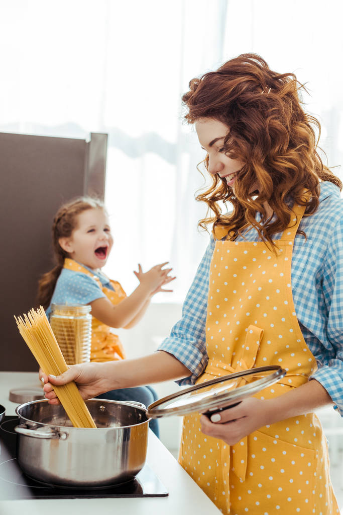 selectieve focus van moeder in polka dot gele schort zetten rauwe spaghetti in pot terwijl opgewonden dochter lachen op achtergrond - Foto, afbeelding