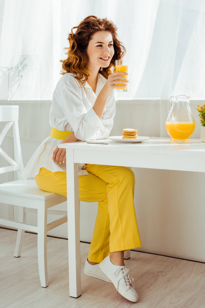 bela mulher feliz sentada à mesa e beber suco de laranja enquanto toma café da manhã
 - Foto, Imagem