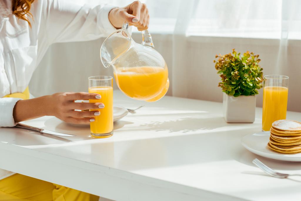 przycięte widok kobiety siedzi przy stole i odlewania soku pomarańczowego w szklance, podczas gdy śniadanie - Zdjęcie, obraz