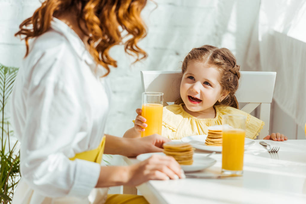 foco seletivo de criança feliz bonito segurando vidro com suco de laranja enquanto toma café da manhã com a mãe
 - Foto, Imagem