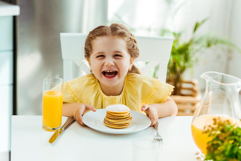 happy excited kid sitting at table, laughing and holding plate with pancakes - Photo, Image