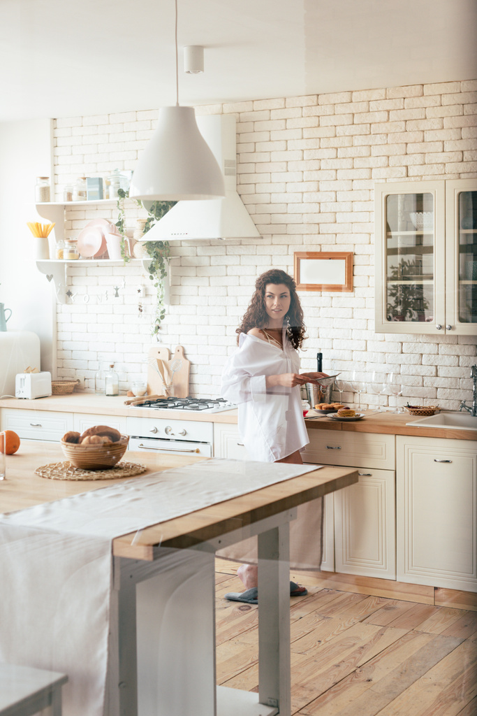 Pretty woman in white shirt looking away while preparing breakfast in kitchen - Photo, Image
