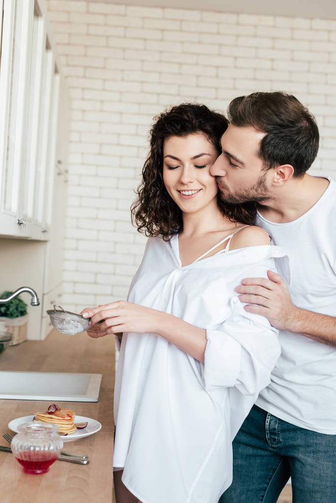 мужчина целует девушку, пока она готовит завтрак на кухне
 - Фото, изображение