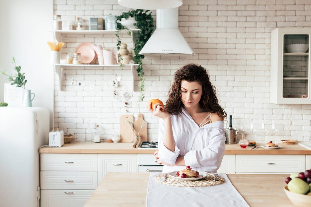 panoramische shot van meisje dat oranje vasthoudt en kijkt naar pannenkoeken in de keuken  - Foto, afbeelding