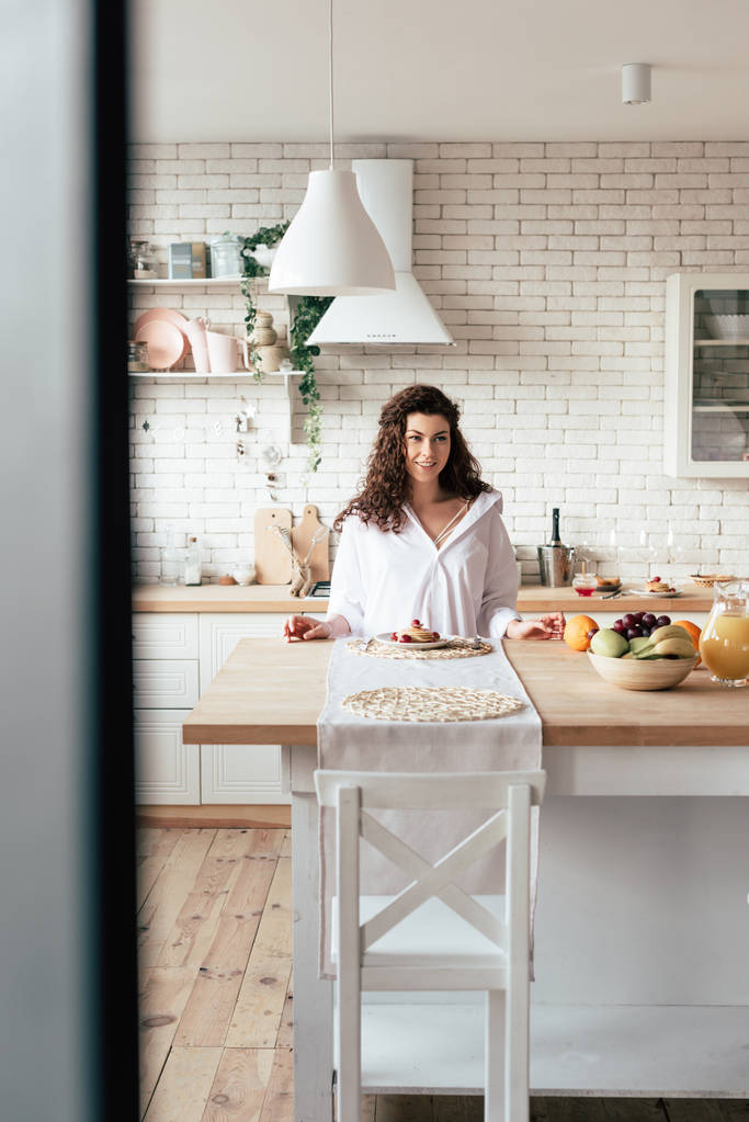 lachende jonge vrouw zittend aan tafel met pannenkoeken en fruit in de keuken - Foto, afbeelding