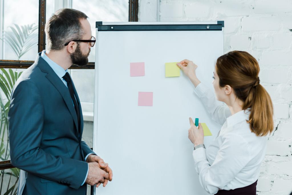 zakenvrouw zetten Sticky Notes op whiteboard terwijl staand met zakenman in glazen - Foto, afbeelding