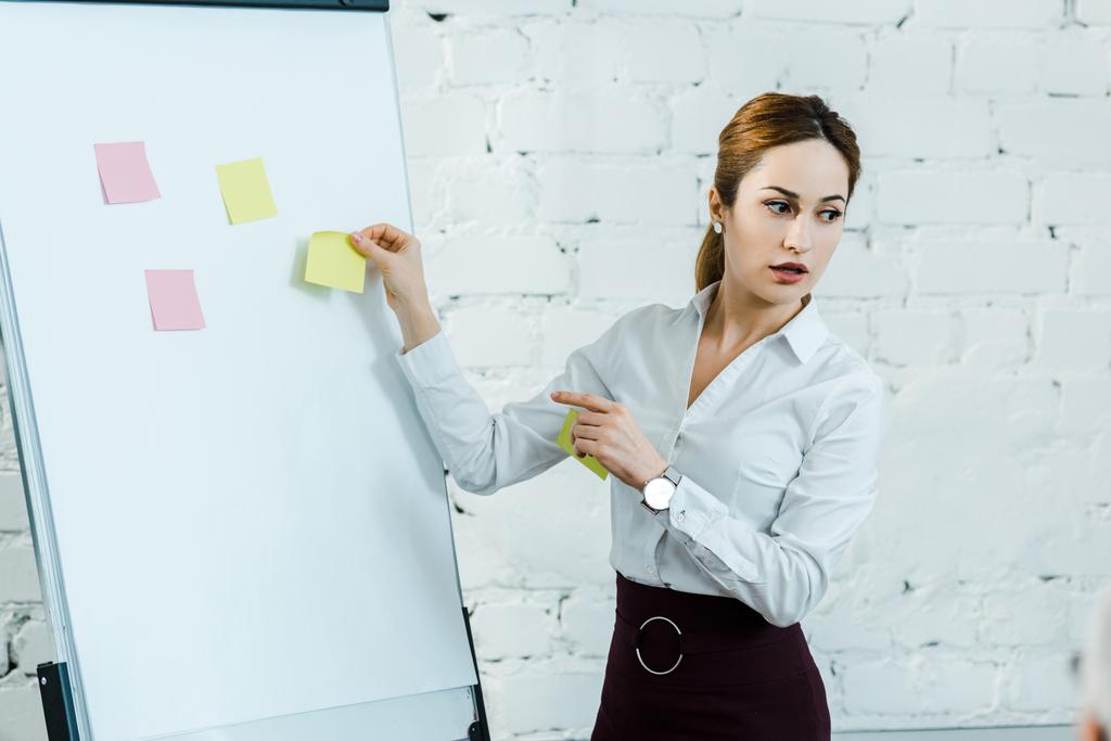 attractive business coach standing near white board and pointing with finger at sticky notes  - Photo, Image
