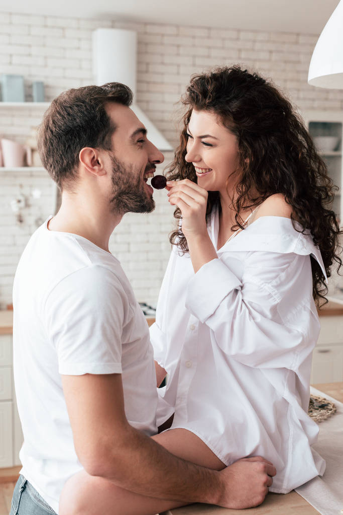 laughing curly woman feeding boyfriend with berry in kitchen - Photo, Image