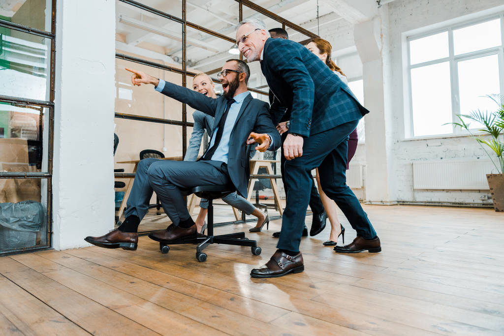 low angle view of cheerful businessman sitting on chair and pointing with finger near multicultural coworkers  - Photo, Image