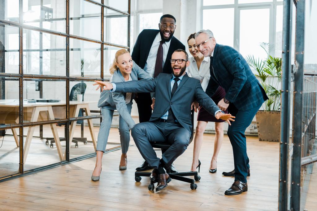 cheerful businessman sitting on chair with outstretched hands near multicultural coworkers  - Photo, Image