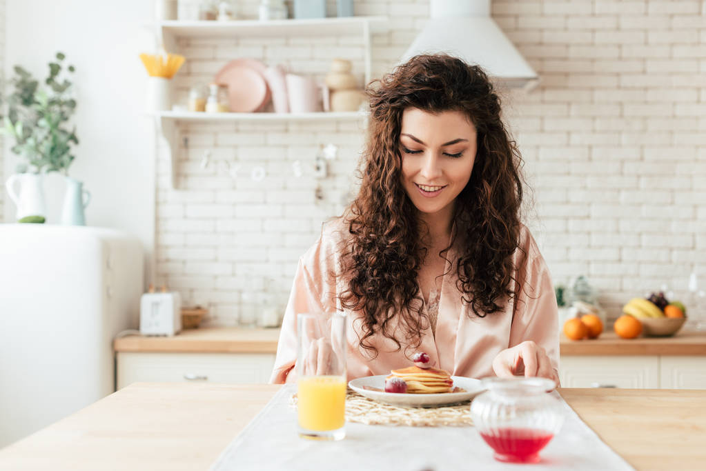 charming curly girl looking at pancakes with smile - Photo, Image