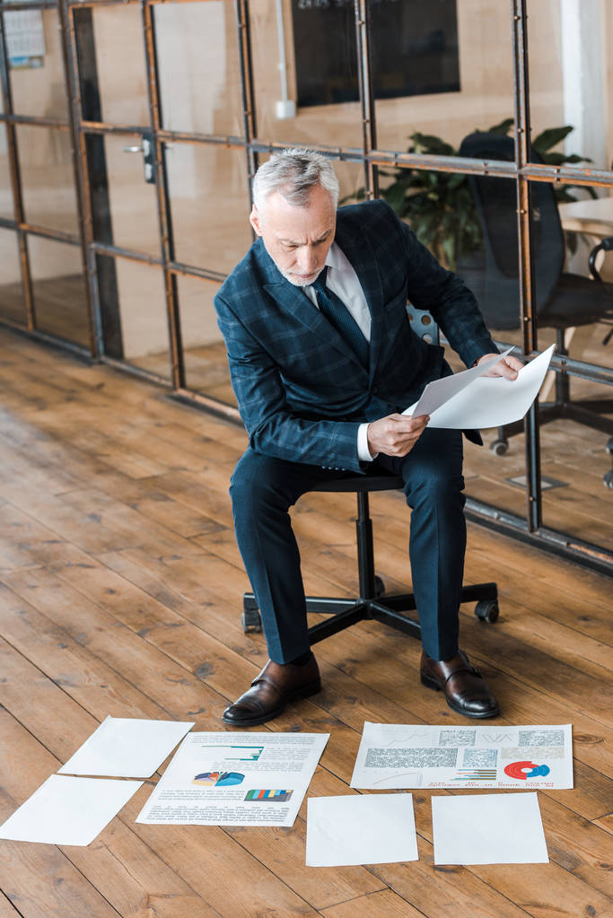 όμορφος επιχειρηματίας που κάθεται στην καρέκλα και κρατάει χαρτιά κοντά σε διαγράμματα και γραφήματα  - Φωτογραφία, εικόνα