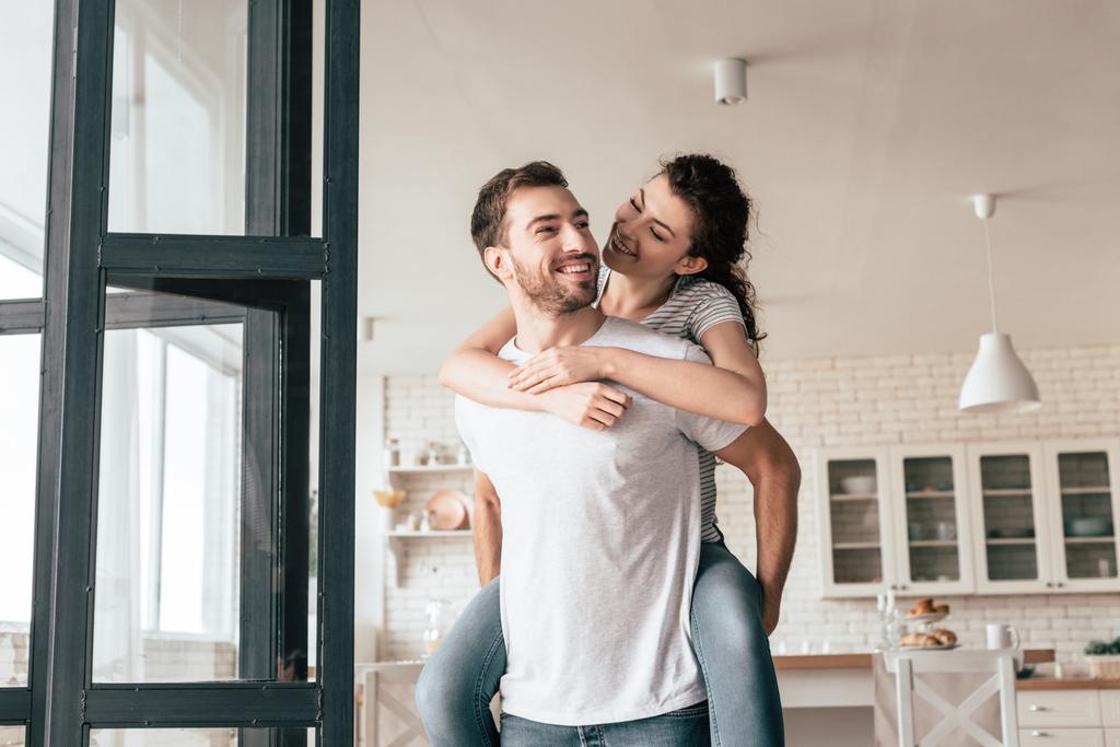 laughing bearded man carrying girlfriend piggyback at home - Photo, Image