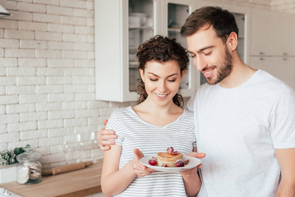 lachende man omarmen vriendin en kijken naar pannenkoeken in de keuken - Foto, afbeelding