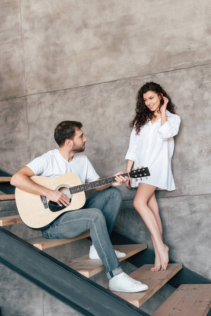 sonriente hombre barbudo sentado en las escaleras y tocando la guitarra acústica a su novia
 - Foto, Imagen
