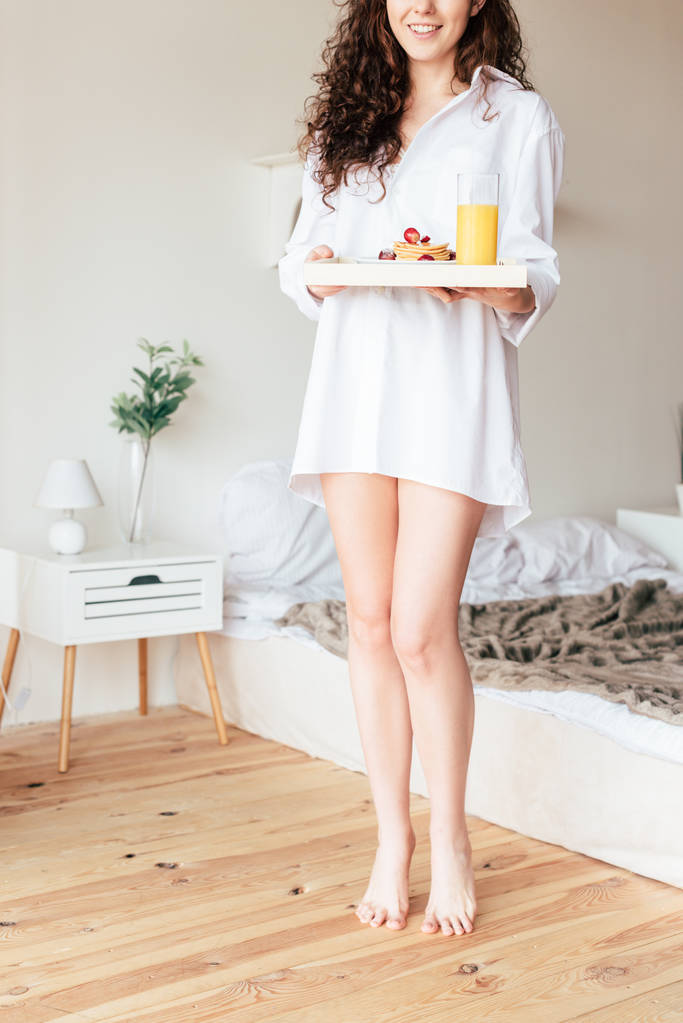 gedeeltelijke weergave van lachende vrouw in shirt met dienblad met ontbijt in de slaapkamer - Foto, afbeelding