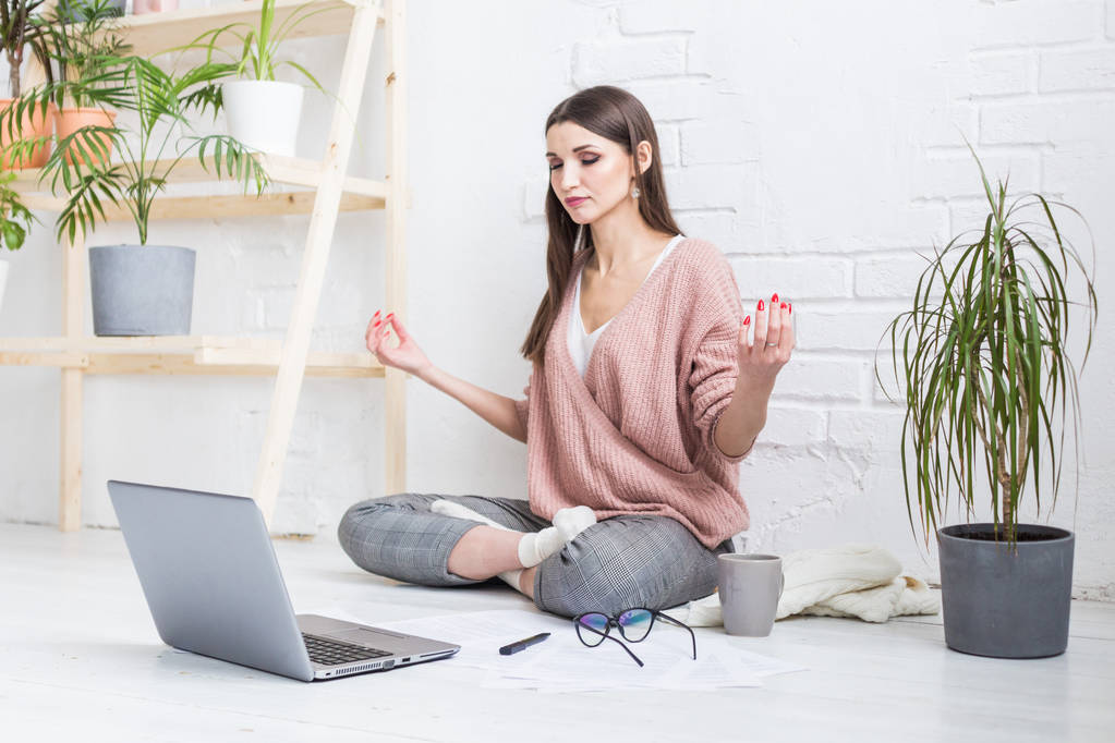 Jovem mulher feliz senta-se no chão em uma pose de ioga em um apartamento brilhante e trabalha atrás de um laptop, uma menina freelancer medita enquanto trabalha. stress de controlo
 - Foto, Imagem