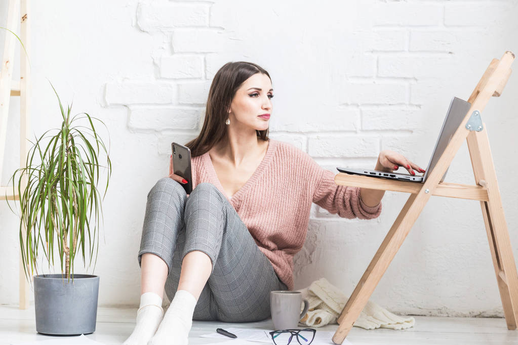 Szczęśliwa kobieta w domowych ubraniach casual siedzi w jasnym wnętrzu i używa telefonu, dziewczyna odpoczywa podczas pracy w laptopie i surfowania po Internecie. Przytulne miejsce pracy freelance, - Zdjęcie, obraz
