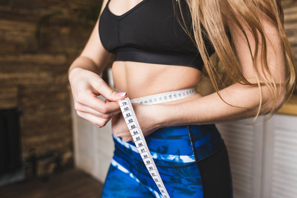 Kaunis nuori nainen urheiluvaatteissa mittaa mitattua vyötärön ympärysmittaa. Urheilu, ruokavalio ja laihtuminen
 - Valokuva, kuva
