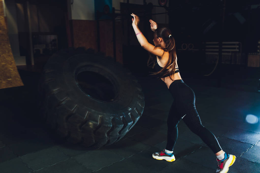 Fitness žena obracející kolo pneumatiky v tělocvičně. Přizpůsobit atletka cvičit s obrovské pneumatiky. Pohled zezadu. Sportovkyně silový trénink. - Fotografie, Obrázek