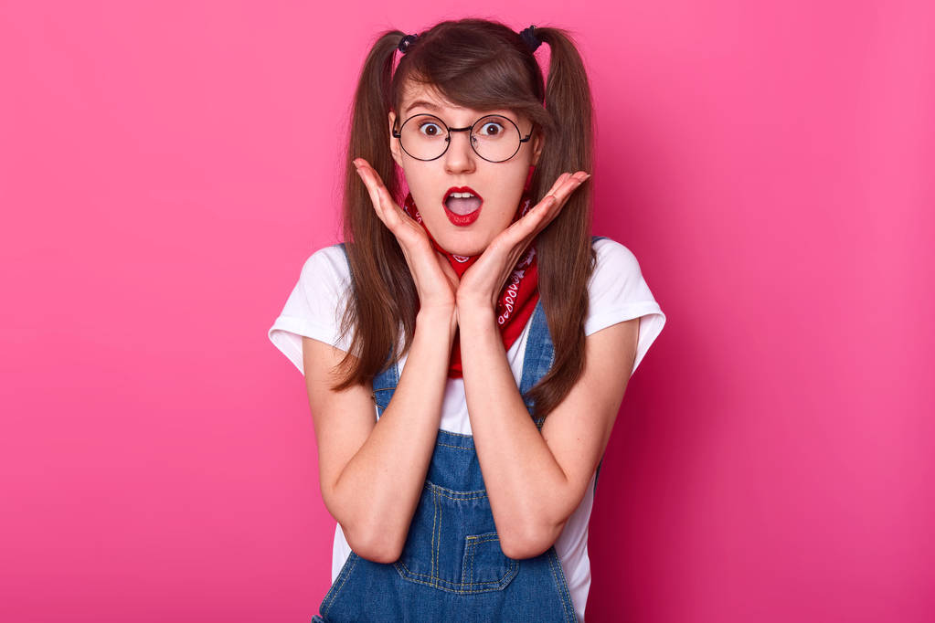 Portrait of astonished emotional girl with pigtails wearing white t shirt, denim overalls and red bandana on neck. Model touches her face with two hands, poses in studio isolated over pink background. - Photo, Image