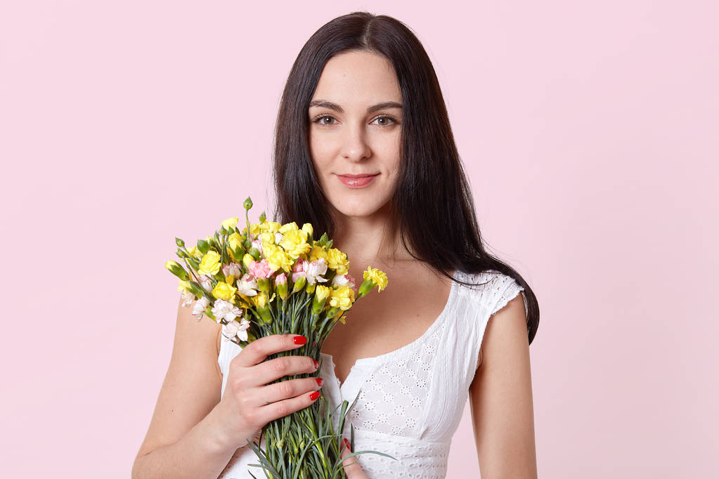 Büyüleyici güzel kadın bir eliyle sarı pembe çiçekler tutar, doğrudan kameraya bakarak, memnun hissediyor. Gülümseyen esmer model beyaz yazlık elbise pozlar. Reklam için kopya alanı. - Fotoğraf, Görsel