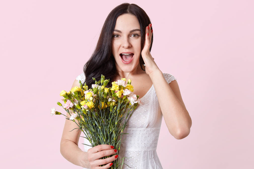 Erstaunt steht die junge Frau vor rosa Hintergrund, öffnet überrascht den Mund, hält einen Strauß Frühlingsblumen in der Hand, sieht glücklich aus. Kopierraum für Werbung. Menschen und Emotionen. - Foto, Bild