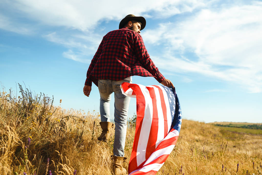 4ο Ιουλίου. Την 4η Ιουλίου. Αμερικανός με την εθνική σημαία. Αμερικάνικη σημαία. Ημέρα της ανεξαρτησίας. Πατριωτική αργία. Ο άντρας φοράει καπέλο, σακίδιο, πουκάμισο και τζιν. Όμορφο φως ηλιοβασίλεμα.  - Φωτογραφία, εικόνα