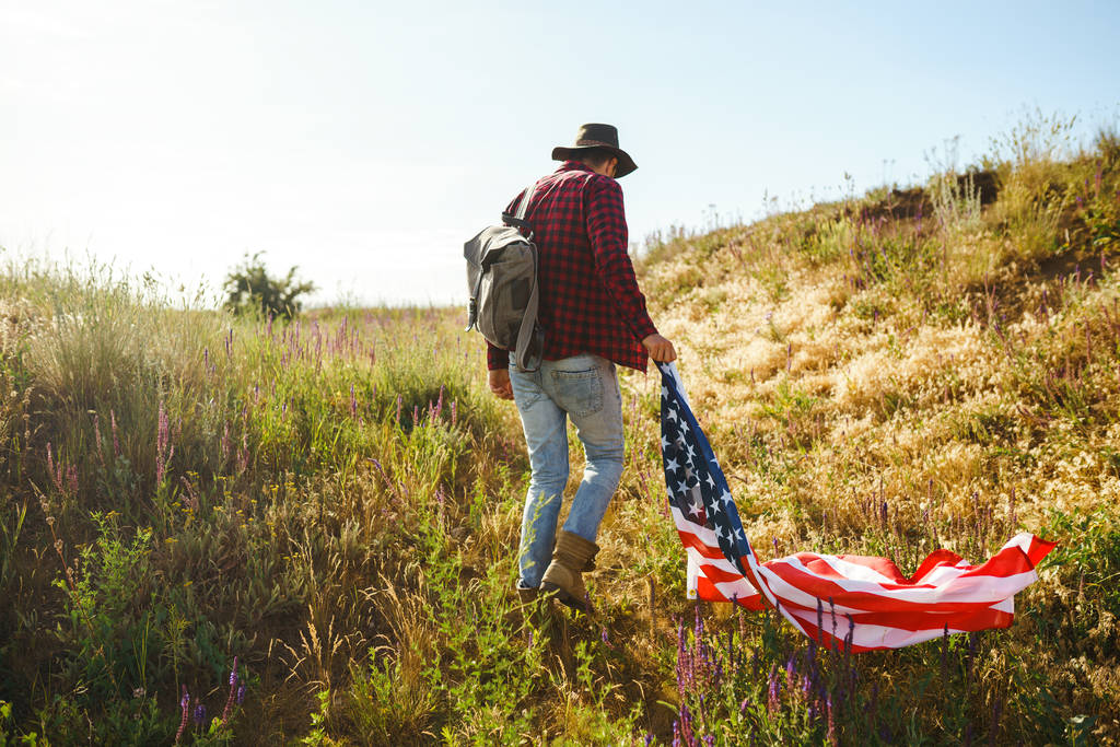 4 de julio. Cuatro de julio. Americano con la bandera nacional. Bandera Americana. Día de la Independencia. Vacaciones patrióticas. El hombre lleva un sombrero, una mochila, una camisa y vaqueros. Hermosa luz de puesta del sol
.  - Foto, imagen