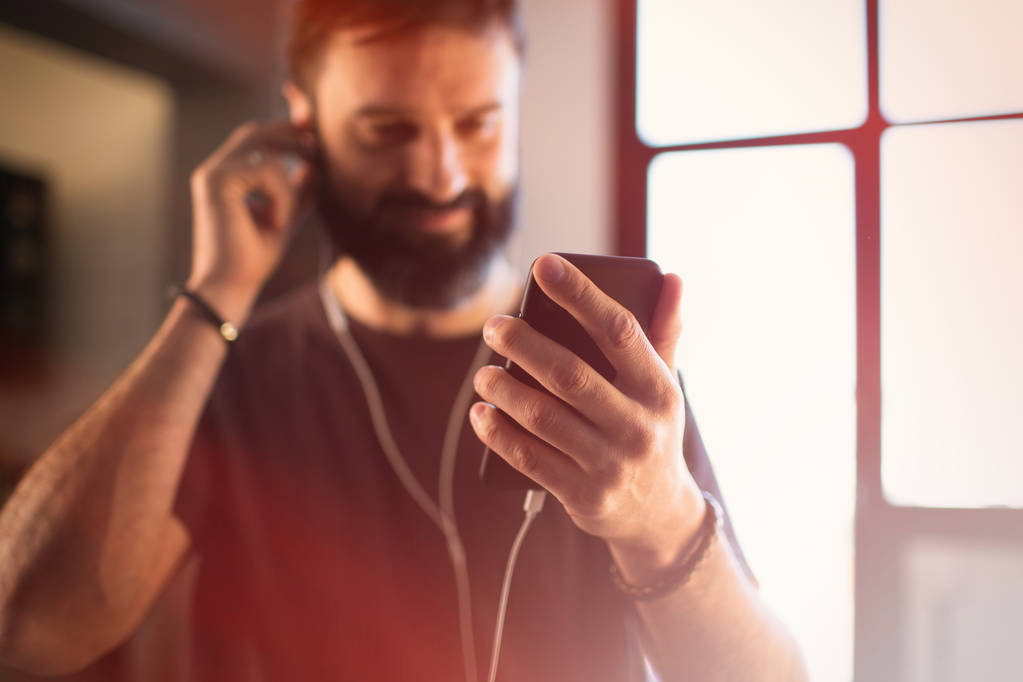 Tipo barbudo confiado que usa una camiseta gris casual escuchando música en los auriculares, comprobando las redes sociales en el teléfono inteligente teniendo feliz en casa
 - Foto, Imagen