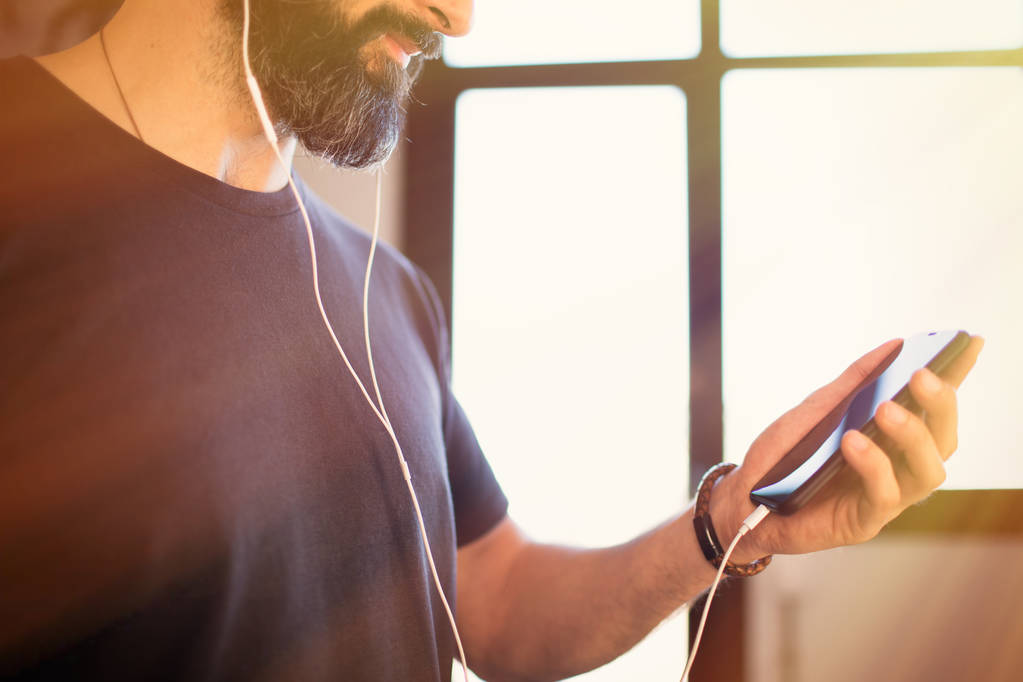 Mosolygó szakállas fickó visel alkalmi szürke póló hallgat zenét fülhallgató, ellenőrzés a szociális hálózatok a mobil telefon otthon - Fotó, kép